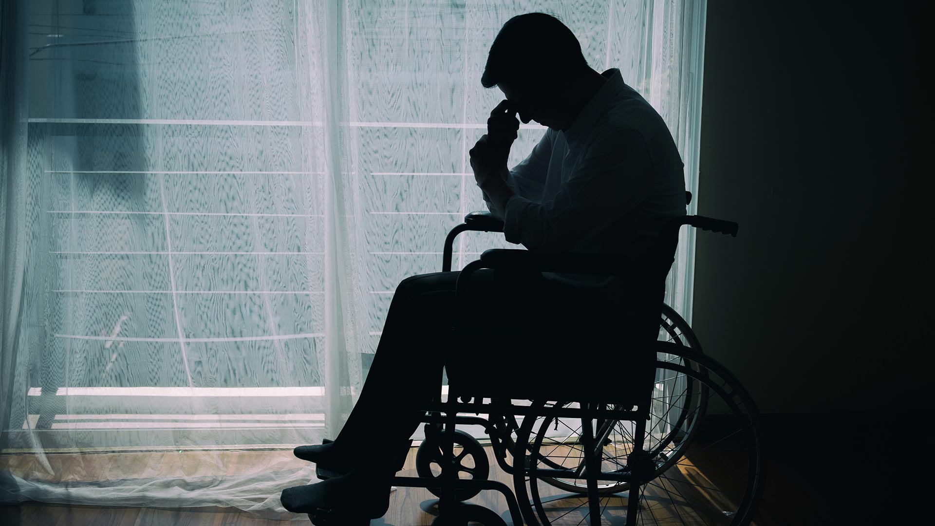 Side profile image of a NDIS Participant in wheel chair silhouetted by window holding head up with hand