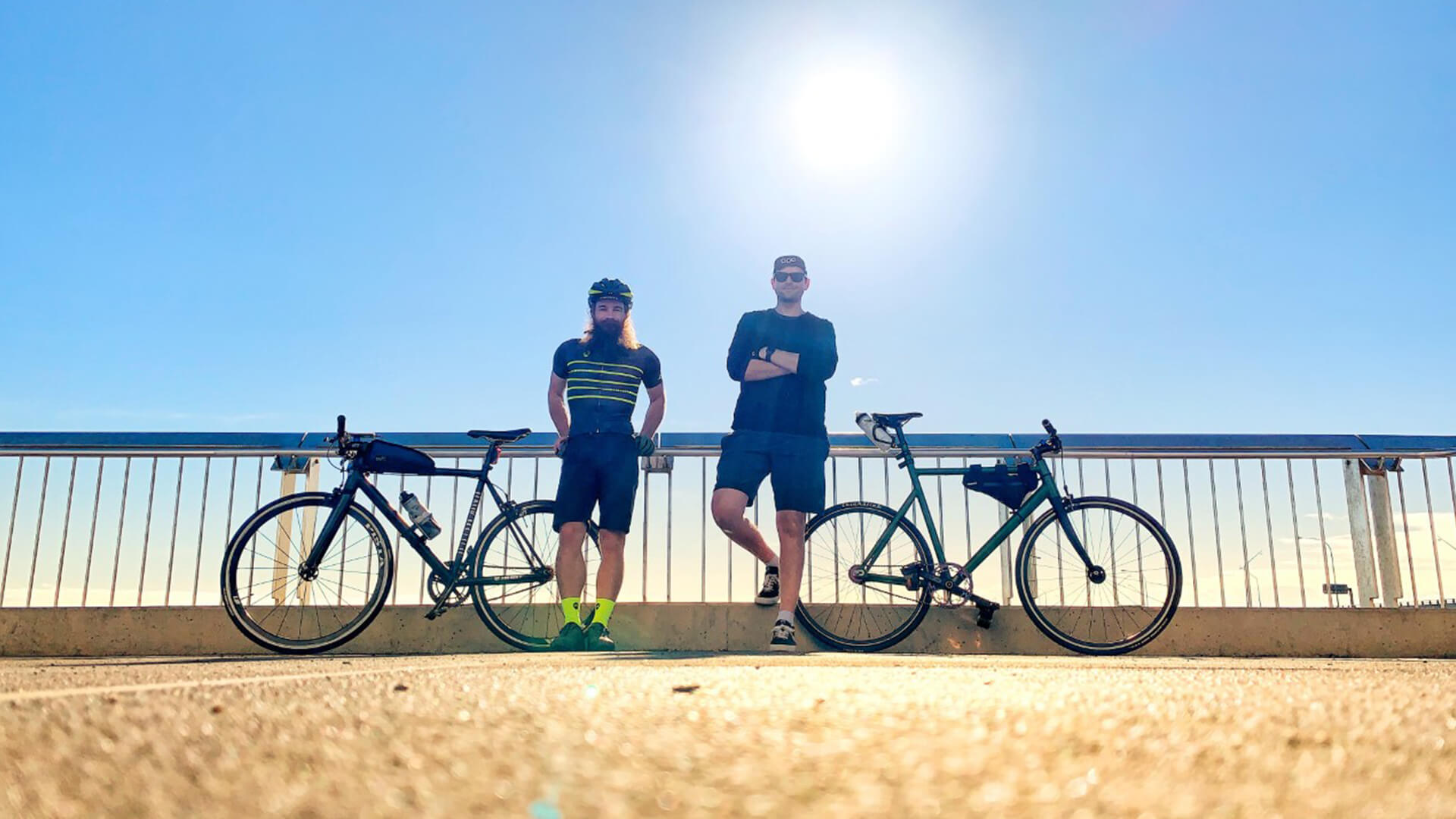 Two MS Brissy to the Bay participants stand side by side against a fence next to their bikes