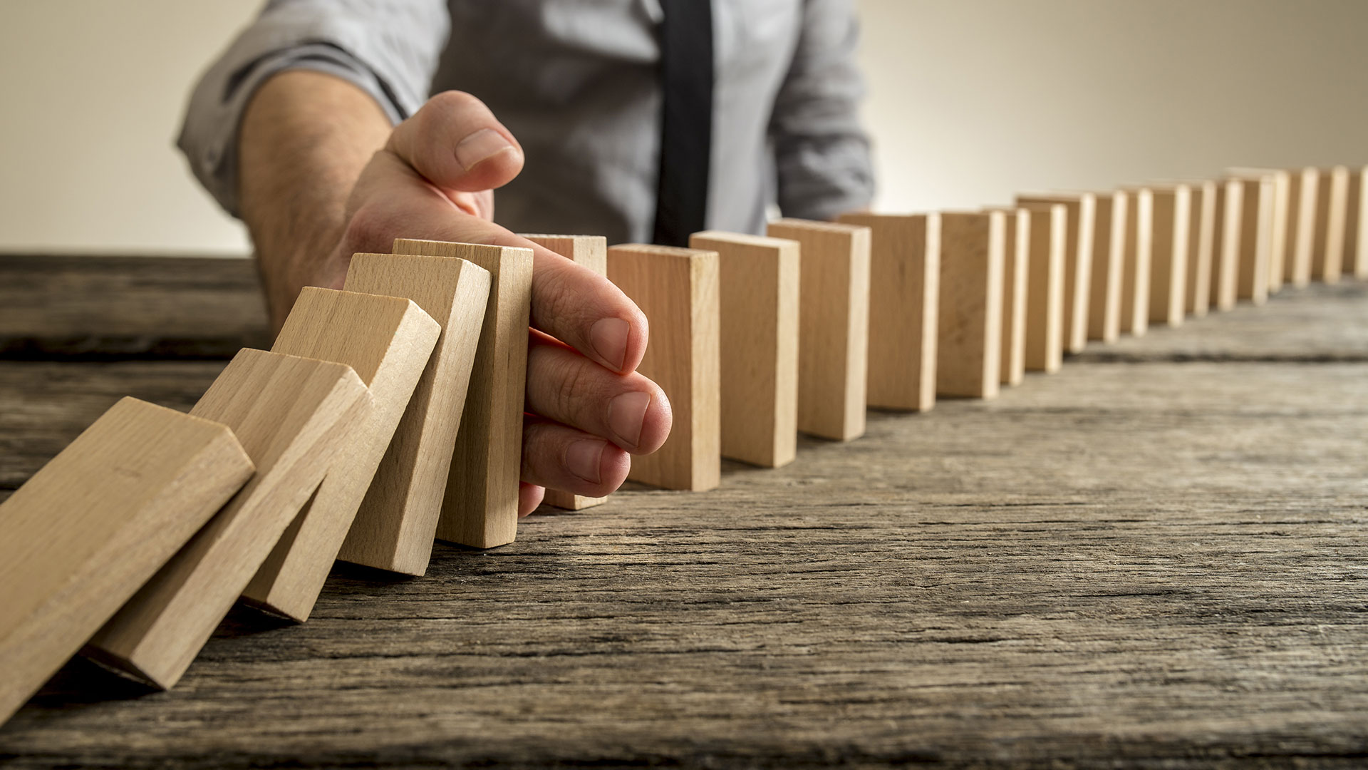 Why are risk assessments so important? A man's hand stops a domino effect on a wooden table