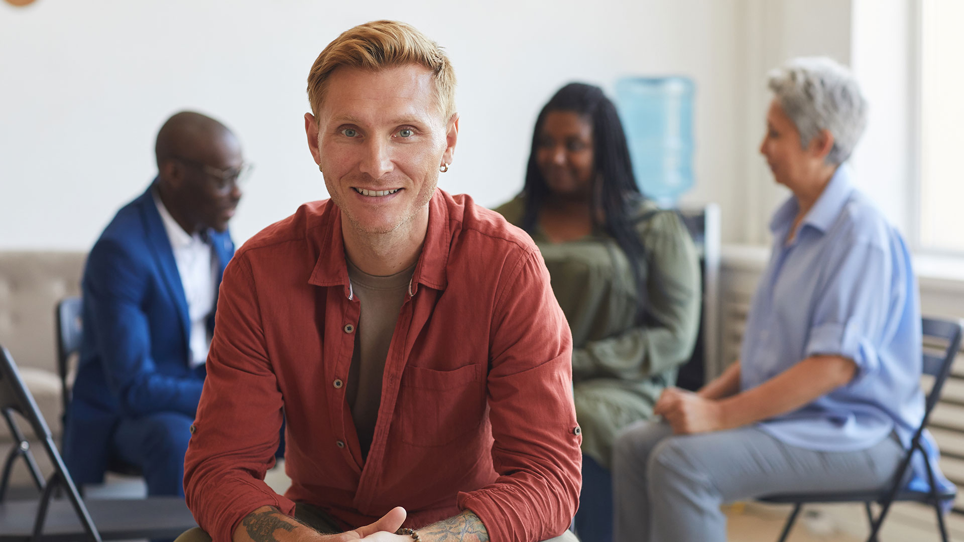 Mental health first aid | A tatooed man sits smiling facing the camera with support group talking in the background