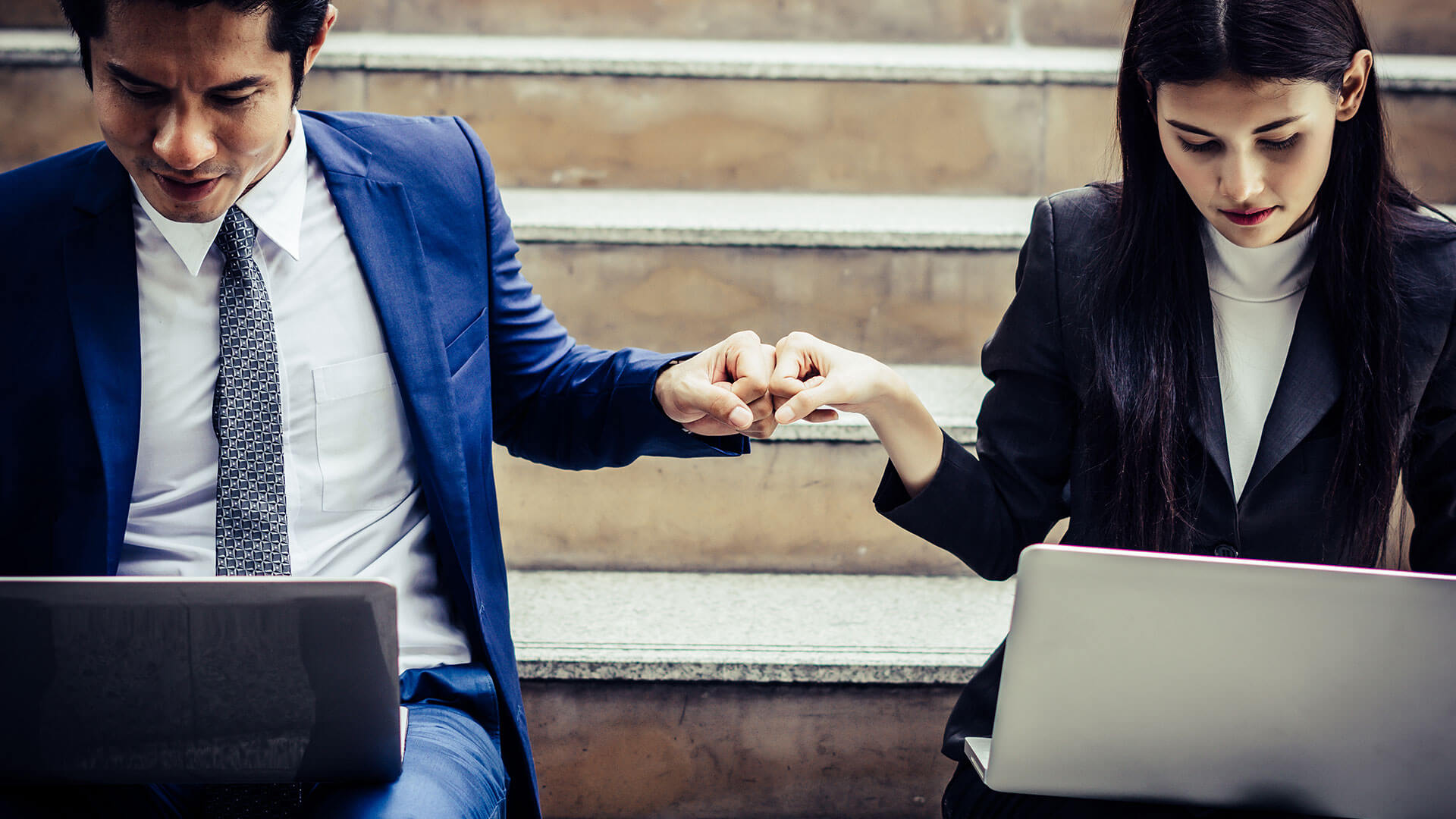 Two young business people sit on stairs with laptops fist bumping. This image represents residential aged care and NDIS providers working together